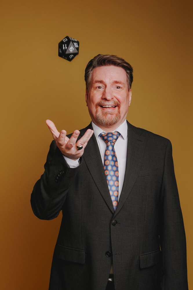 Ein Mann im Anzug lächlet in die Kamera und wirft dabei mit seiner rechten Hand ein größeren 20-seitigen Würfel in die Luft